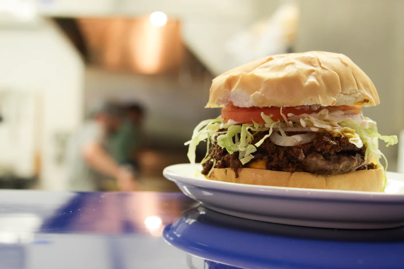 A burger topped with chili, cheese, onion, lettuce and tomato, with the kitchen behind it