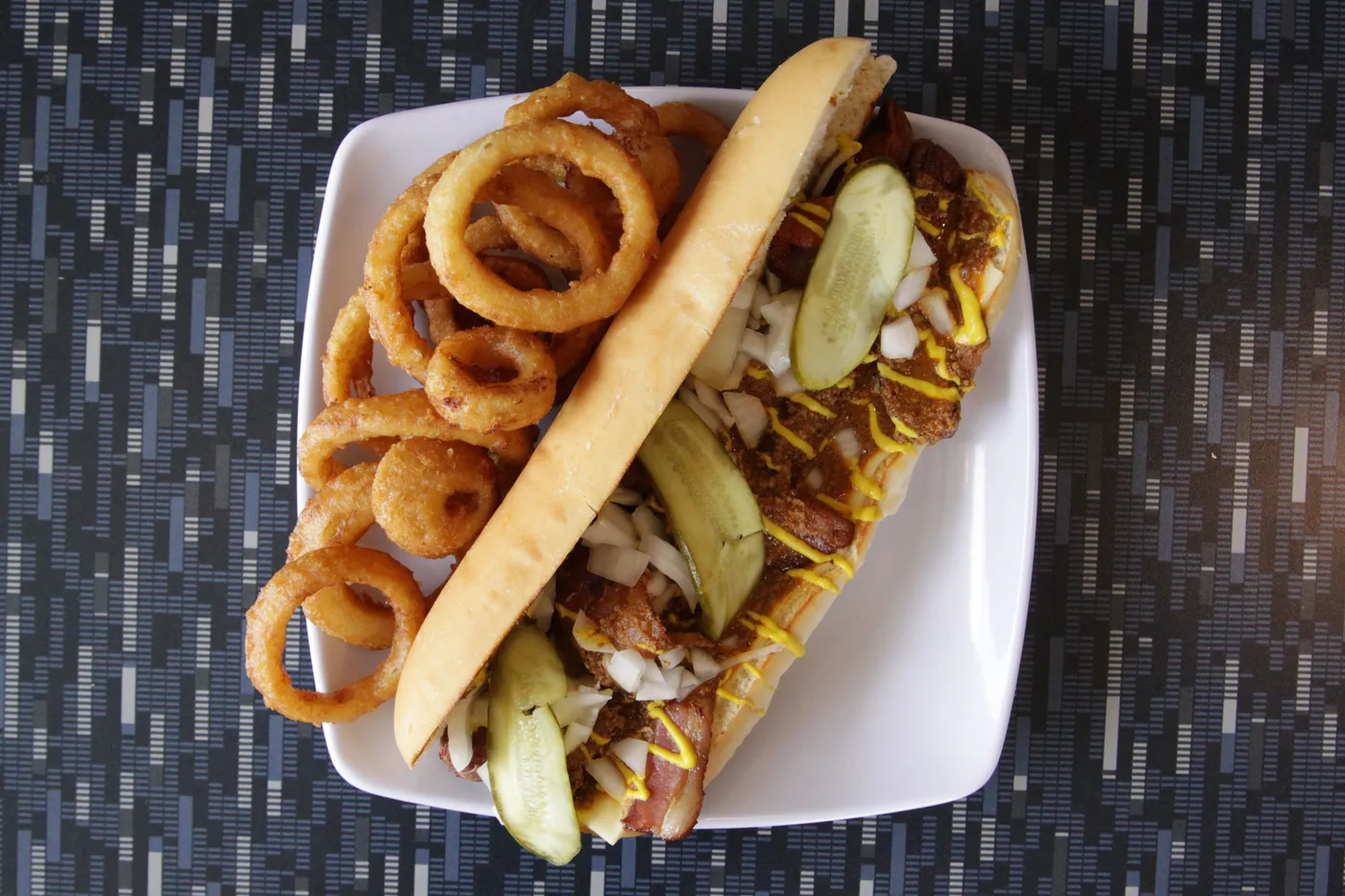 A foot long hot dog topped with chili, onion, mustard and a pickle, with onion rings