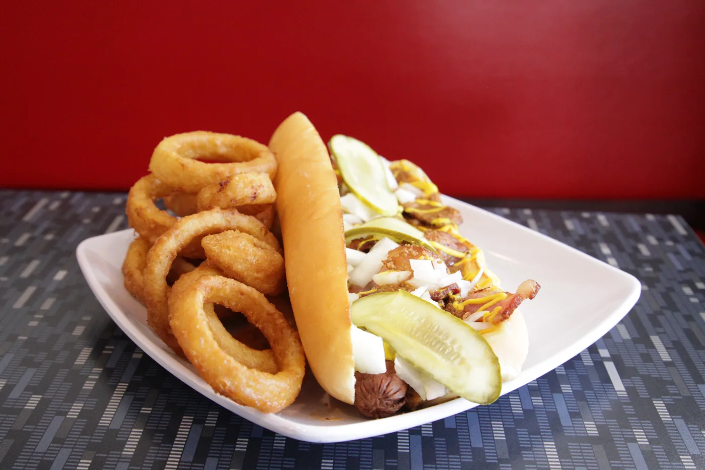 A foot long hot dog with a pickle, beside a pile of onion rings